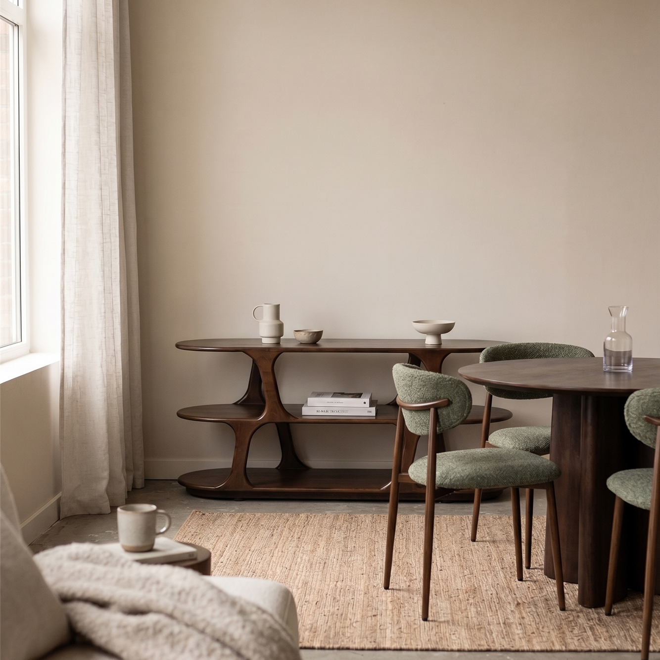 A styled interior room scene showing a mid-century modern wooden sideboard/console table, a round dining table with green upholstered chairs, a woven jute rug, and a light beige sofa in a softly lit room.
