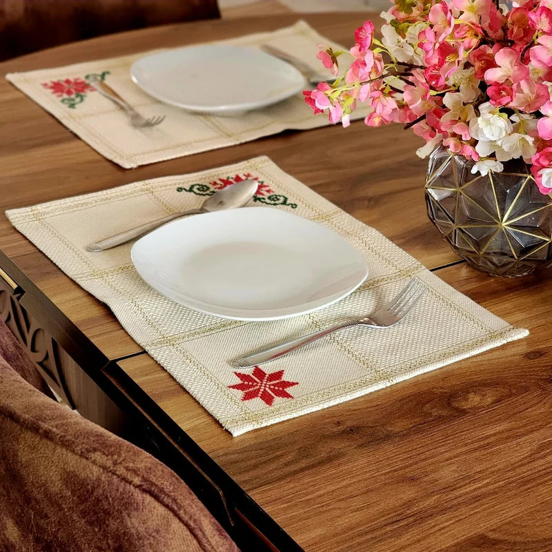 Two woven beige placemats with embroidered red and green floral motifs are set on a wooden dining table, each styled with a white plate, silver fork, and knife. A vase of pink flowers sits on the right side of the table.