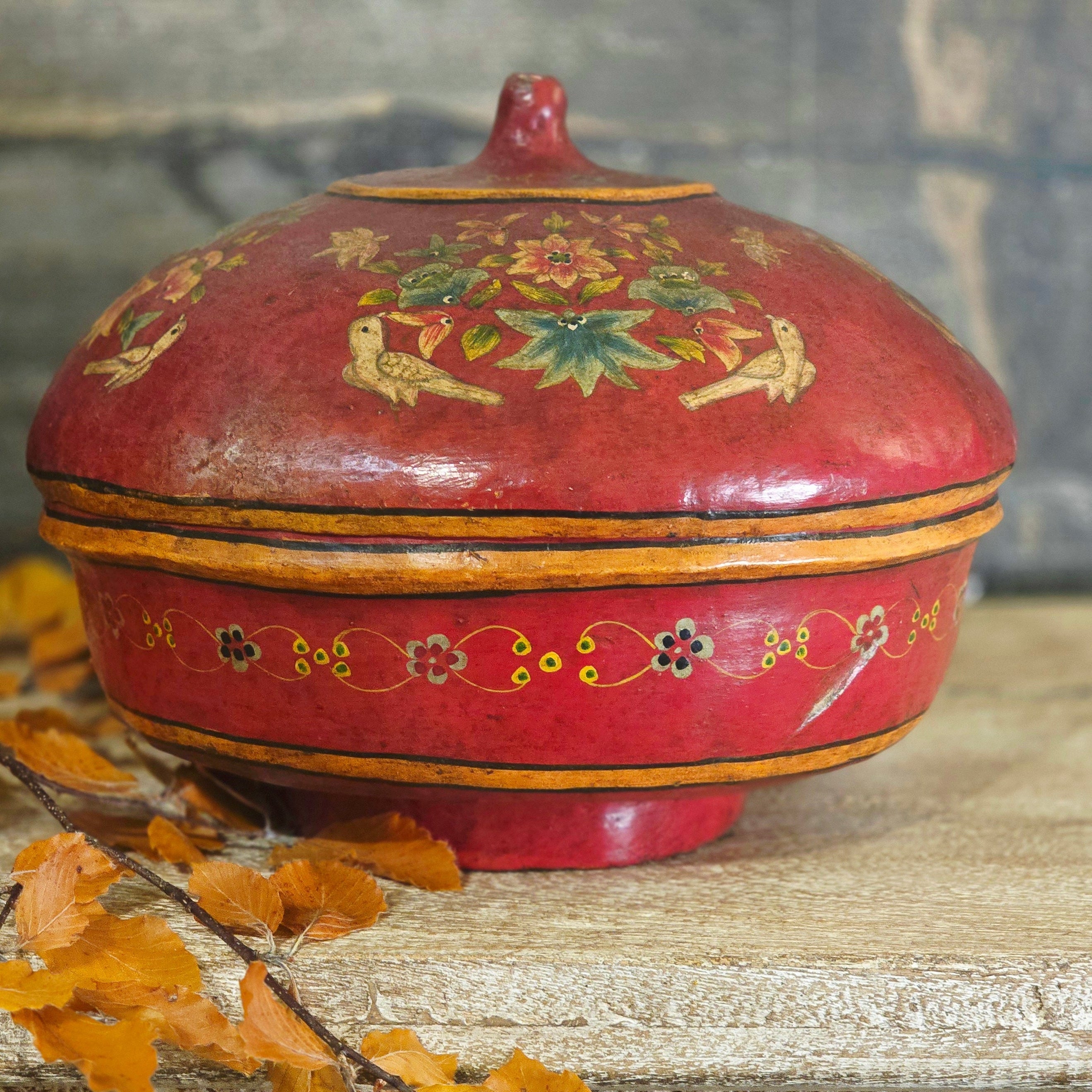A round red lacquered wooden lidded box with hand-painted floral and bird motifs, displayed on a distressed wooden surface with dried autumn leaves in the foreground.